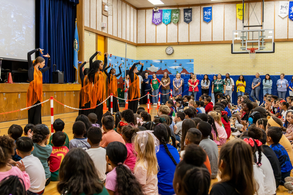 Student dancers wearing black outfits with bright orange fringe perform in front of a large audience of seated elementary students inside the school gymnasium.
