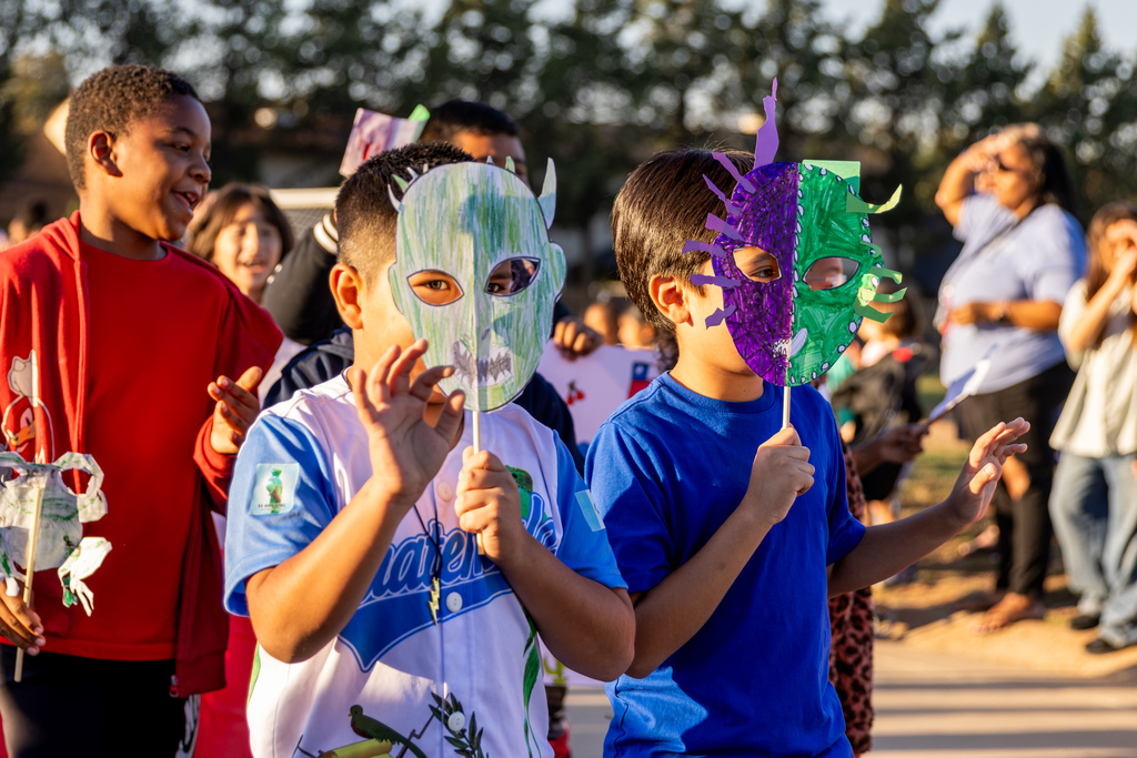 Two students hold colorful handmade masks in front of their faces while walking in a parade, surrounded by classmates enjoying the celebration.