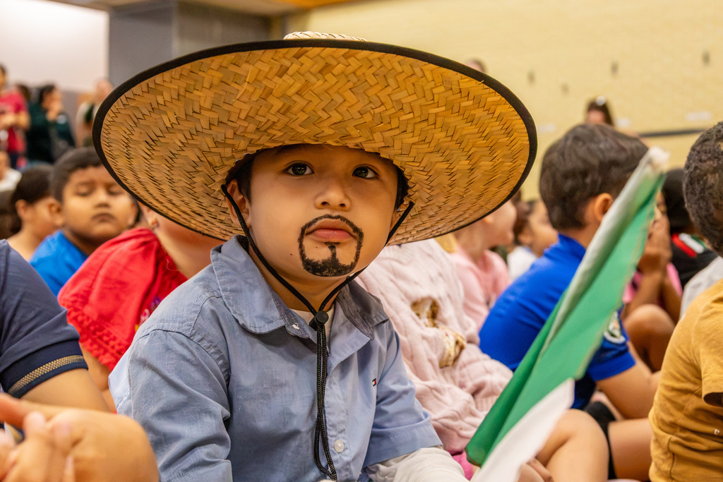A student wearing a large woven sombrero and a drawn-on mustache and beard looks toward the camera while seated among classmates during the celebration.