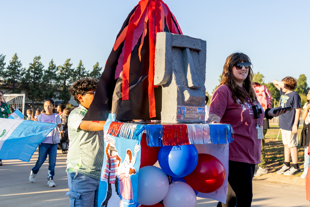 A student and staff member walk alongside a decorated parade float featuring a replica moai statue representing Easter Island, Chile, with red, white, and blue balloons.
