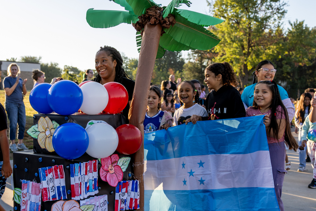 Smiling students carry a Honduran flag alongside a handmade tropical-themed float decorated with a palm tree, flowers, and red, white, and blue balloons.