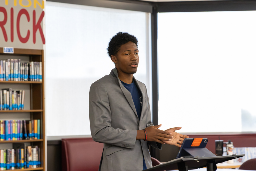 A Putnam City North student in a gray blazer stands behind a lectern, gesturing with his hands as he presents an argument during a mock trial of the Mongol Empire. Library bookshelves and windows fill the background.