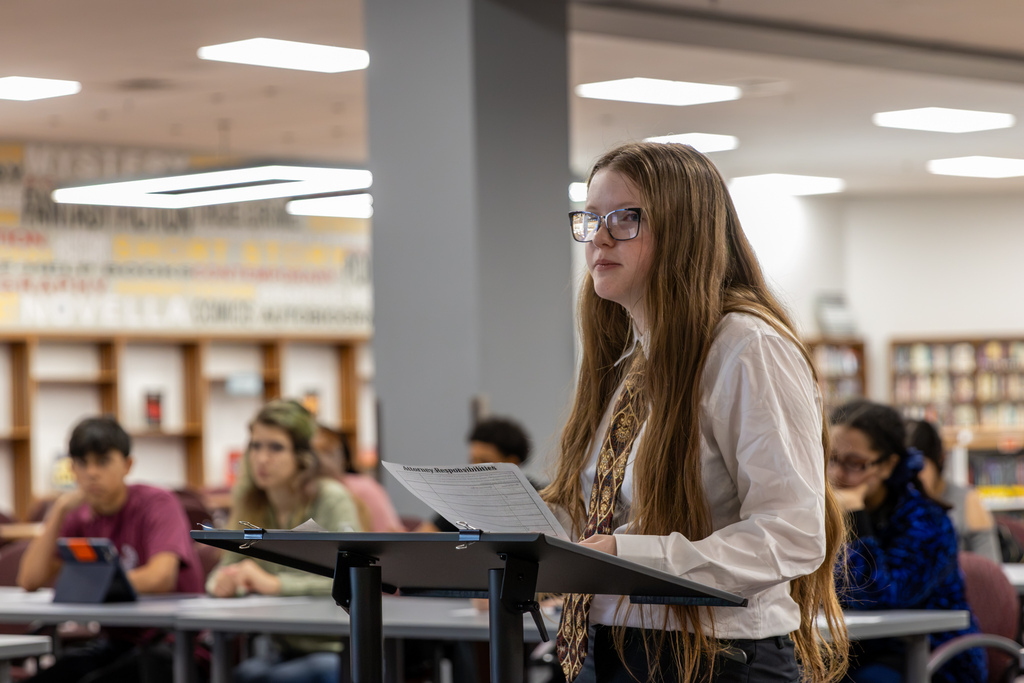 A Putnam City North student wearing glasses, a white dress shirt, and a patterned tie stands behind a lectern holding notes during an AP World History mock trial. Classmates listen in the background.