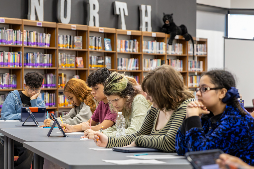 Putnam City North students sit in a row at library tables, reviewing documents, writing notes, and using tablets as they prepare their arguments during a mock trial of the Mongol Empire.