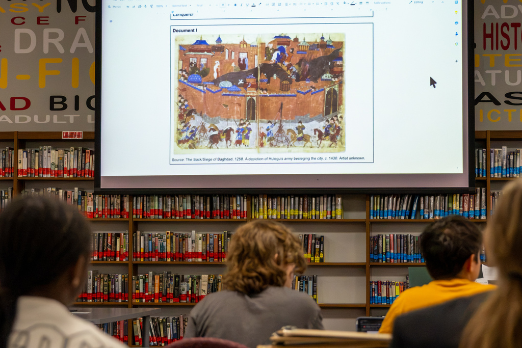 A projected historical illustration of the 1258 Siege of Baghdad appears on a screen in the Putnam City North library as students examine evidence during their mock trial of the Mongol Empire.