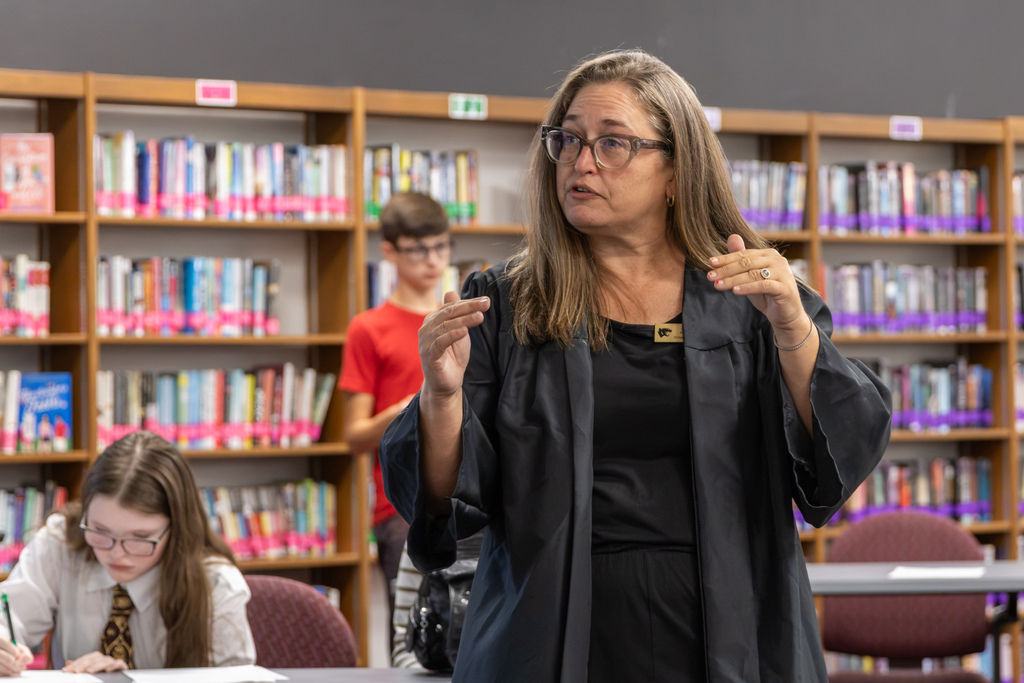 A Putnam City North teacher wearing glasses and a black blazer gestures with both hands while speaking to students during an AP World History mock trial in the school library.