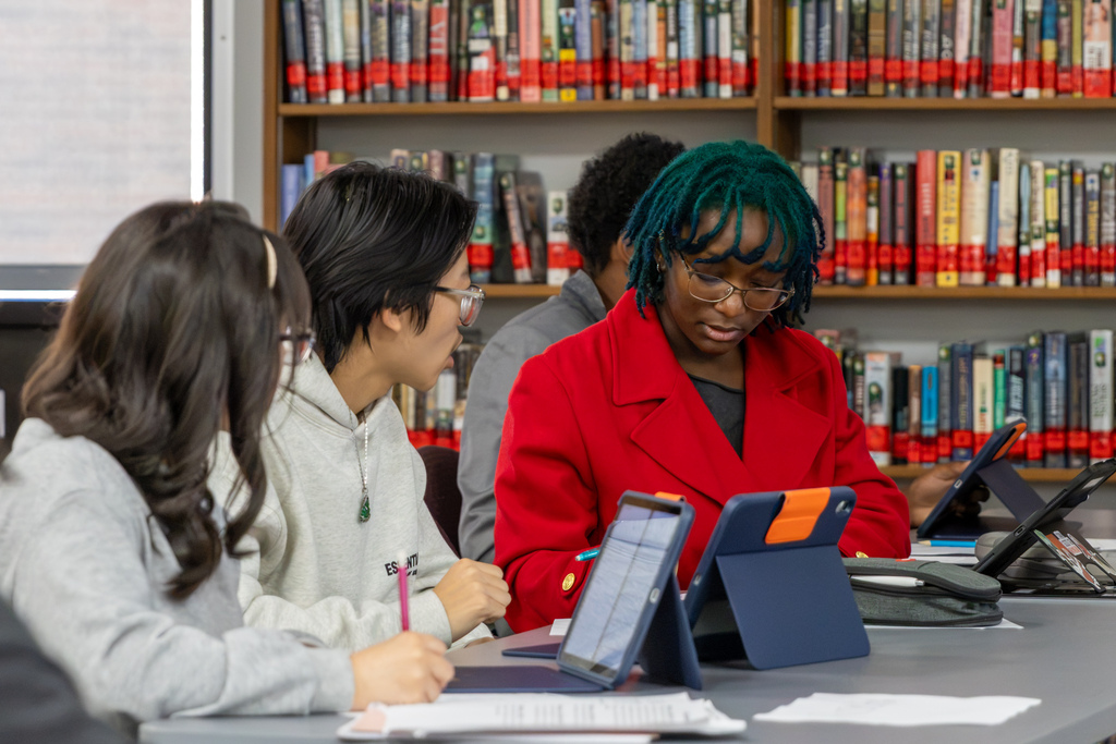 A Putnam City North student wearing a bright red blazer and glasses reviews information on a tablet while classmates beside them consult notes during an AP World History mock trial.