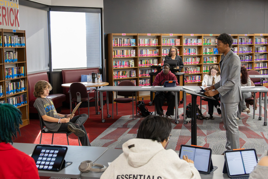 A Putnam City North student in a gray suit stands at a lectern presenting an argument during a mock trial in the school library. Other students sit at tables with notes and tablets, while a student across the room holds a clipboard.