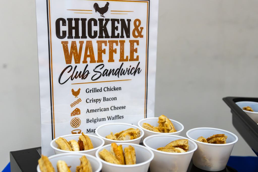 Small white cups containing bite-sized chicken and waffle club sandwich samples sit in front of a display sign listing grilled chicken, crispy bacon, American cheese, Belgian waffles, and maple brown sugar mustard as ingredients.