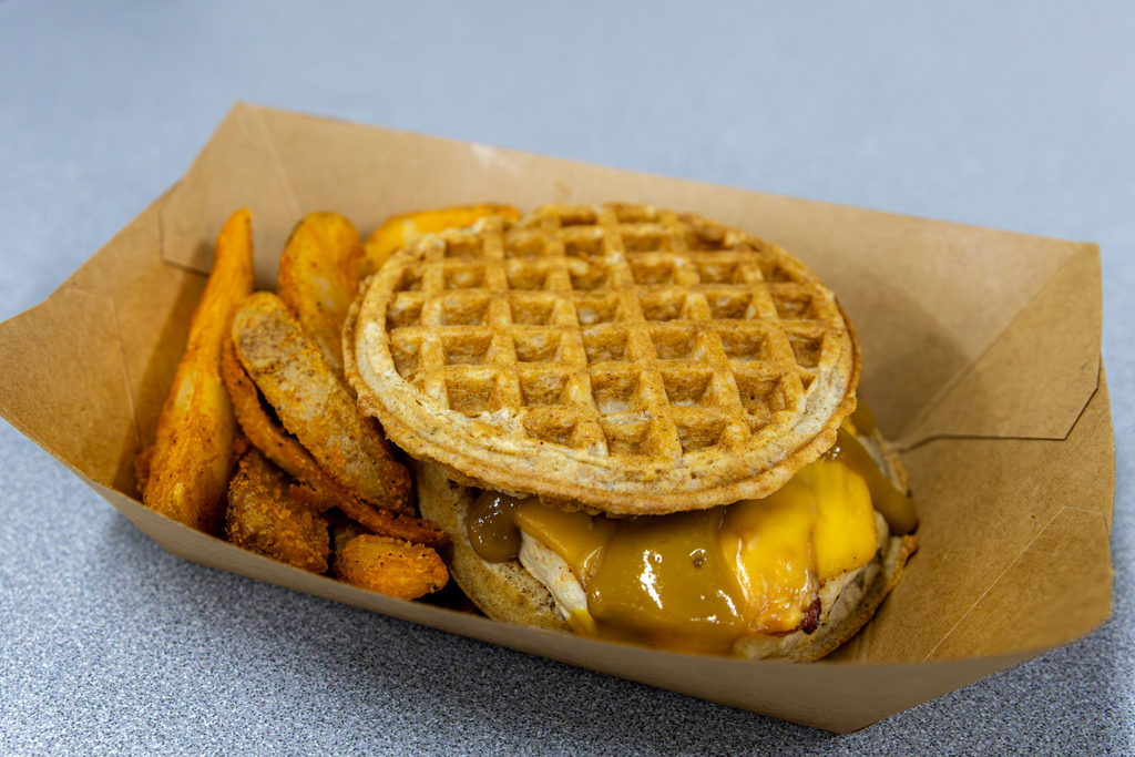 Close-up of a chicken and waffle club sandwich served with seasoned potato wedges in a cardboard tray. The sandwich features grilled chicken, melted cheese, and maple brown sugar mustard between two Belgian waffles.