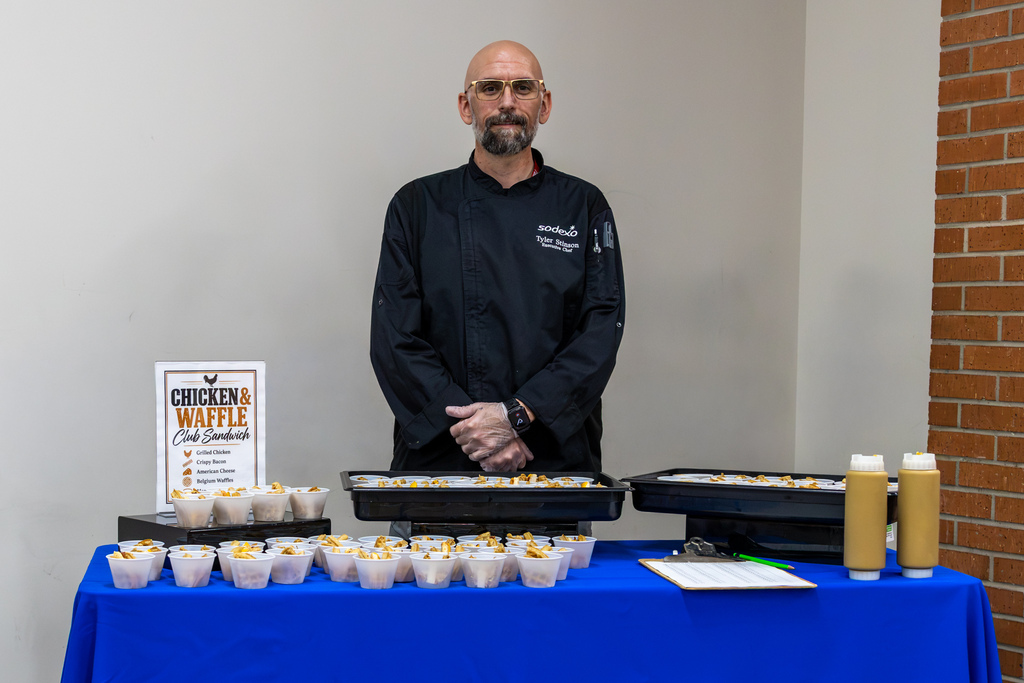 Sodexo Executive Chef Tyler Stinson stands behind a table filled with sample cups of chicken and waffle club sandwiches at Putnam City High. A menu sign and bottles of sauce sit alongside trays of samples.