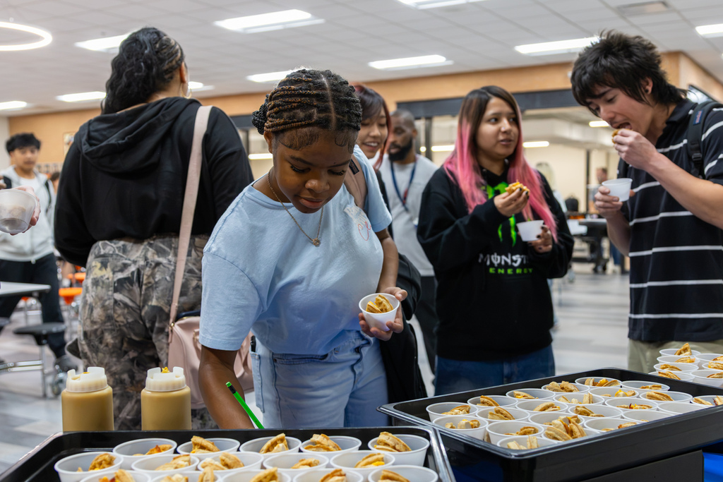 Putnam City High students sample chicken and waffle club sandwiches in the cafeteria. One student reaches toward a feedback sheet while others taste their samples beside trays of food.