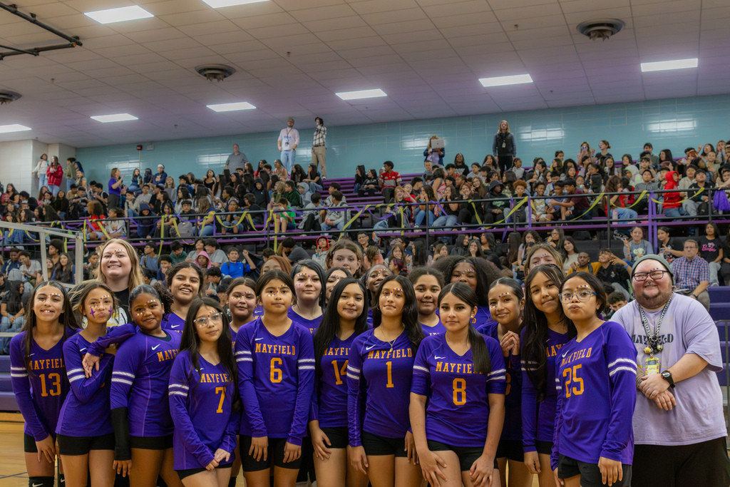 Mayfield Middle School volleyball players and staff pose together in purple and gold in front of a packed gymnasium. Students wearing purple and yellow face decorations stand shoulder to shoulder, with classmates filling the bleachers behind them.
