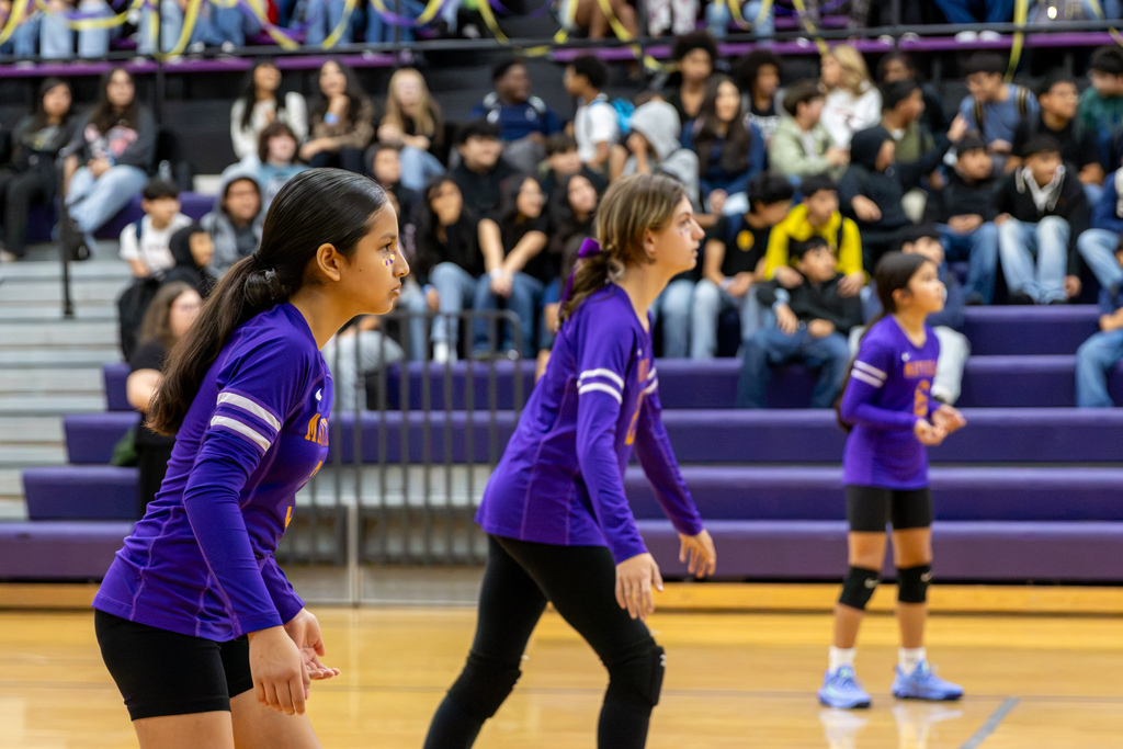 Three Mayfield Middle School volleyball players in purple uniforms stand in ready positions during a scrimmage, with students watching from the bleachers in the background.