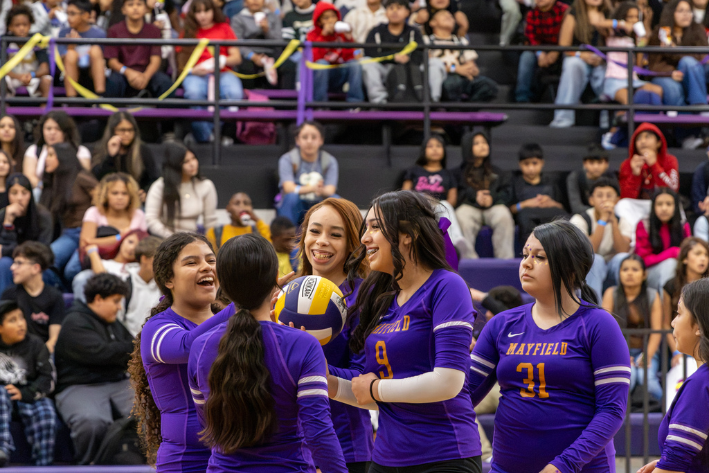 Mayfield Middle School volleyball players in purple uniforms gather around a volleyball, smiling and laughing together during their end-of-season scrimmage as classmates watch from the stands.