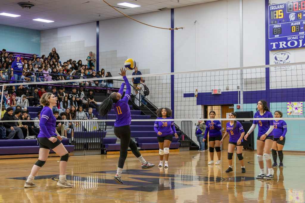 A Mayfield Middle School volleyball player wearing jersey number 11 reaches up to hit the ball over the net during a varsity versus JV scrimmage. Teammates watch the play as a large crowd fills the bleachers.