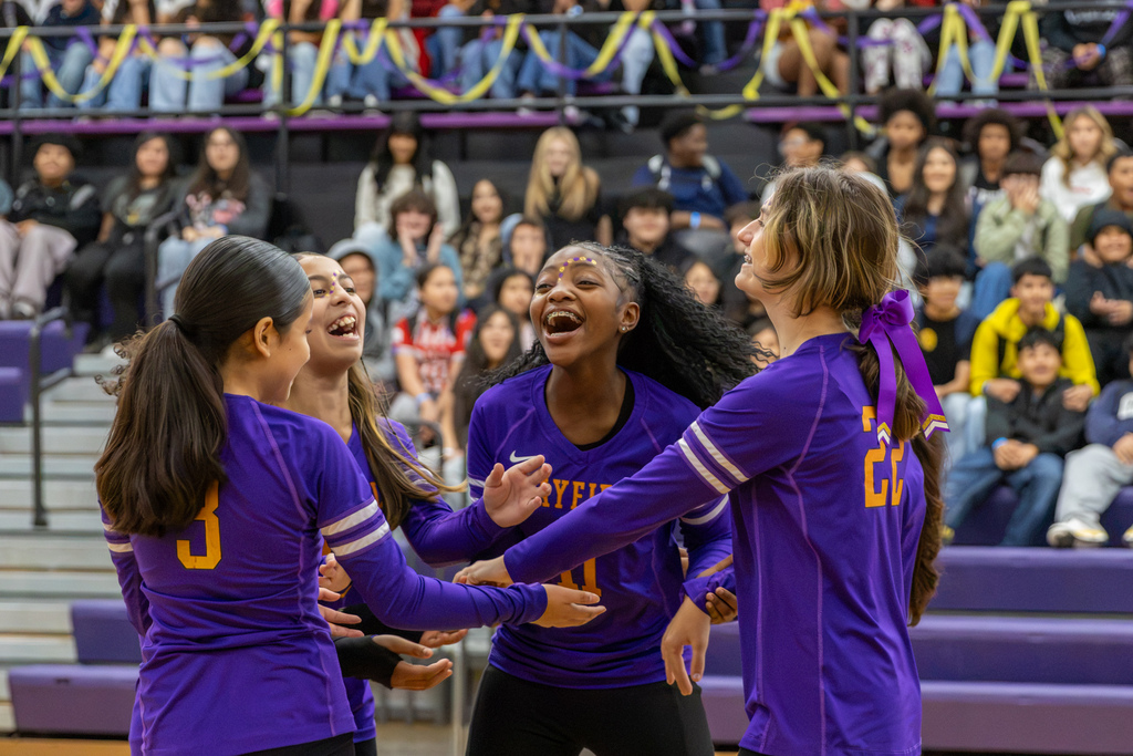 Four Mayfield Middle School volleyball players in purple jerseys laugh and celebrate together on the court, with classmates watching from the packed bleachers behind them.