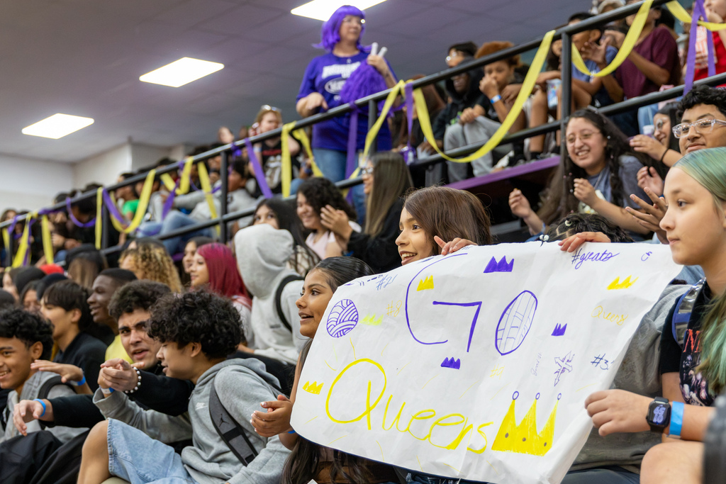 Mayfield Middle School students cheer from crowded gym bleachers decorated with purple and yellow streamers. Two students in the foreground hold a handmade poster reading "GO Queens" with volleyball drawings and crowns.