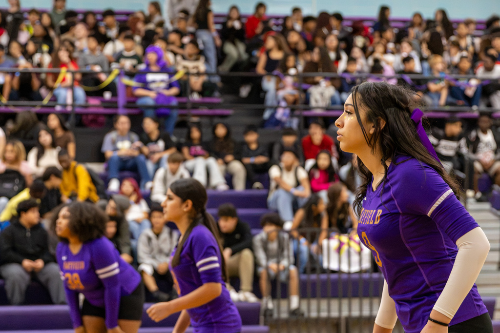 Three Mayfield Middle School volleyball players prepare for the next play on the gym floor, with a player wearing jersey number 9 in the foreground and packed bleachers behind them.