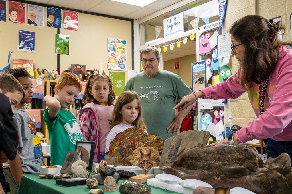 Randy Utt points out fossils displayed on a classroom table as Dennis Elementary students examine the collection, with her husband Dale standing nearby.