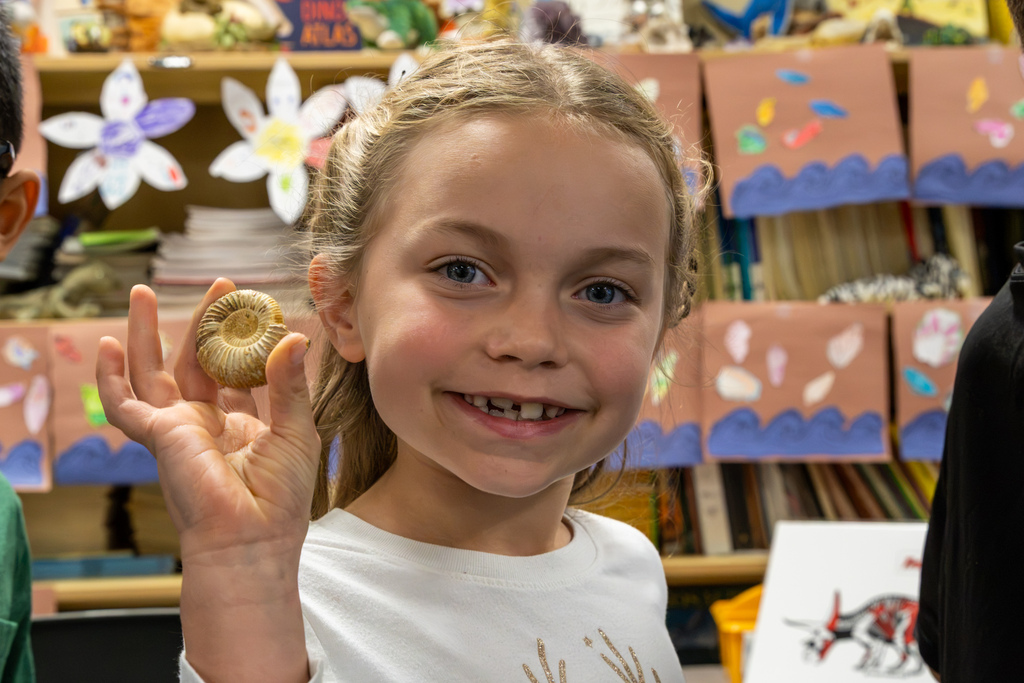 A smiling Dennis Elementary student proudly holds a small spiral-shaped ammonite fossil beside her face.