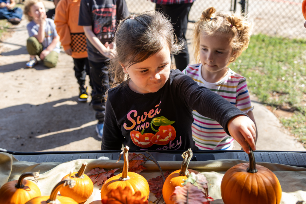 A young girl in a black long-sleeve shirt reaches down to pick up a small pumpkin lined up inside the back of an SUV, with another student watching beside her.