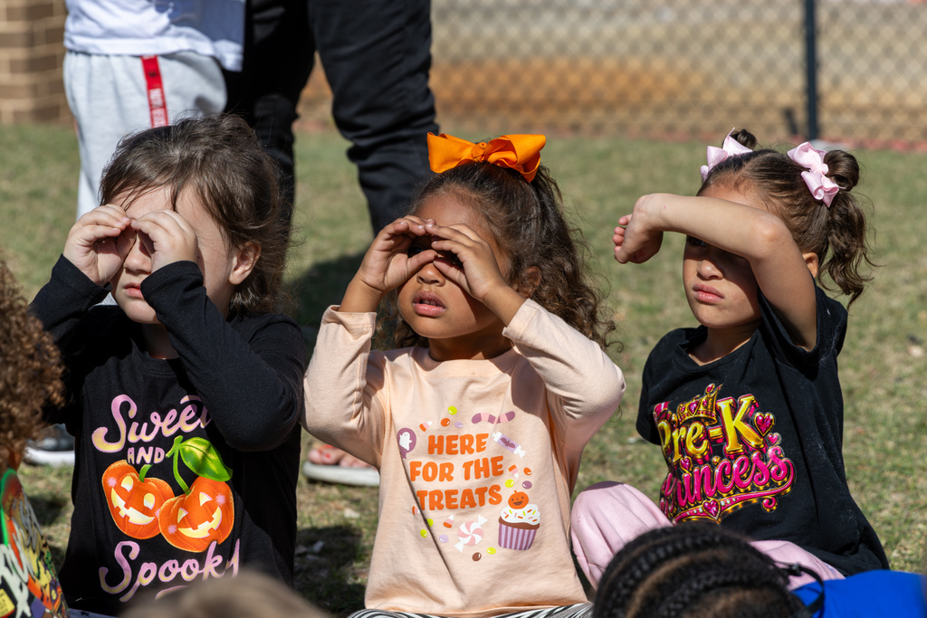 Three young Pre-K students sit on the grass outside, cupping their hands around their eyes like binoculars to peer ahead.