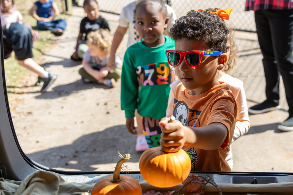 A young boy wearing red sunglasses reaches his hand out toward an open SUV trunk to grab a small pumpkin.