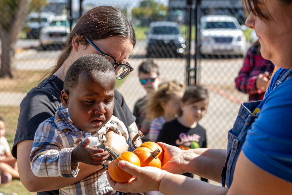 A school staff member holds up two small pumpkins for a young student held in an adult's arms to touch and inspect.