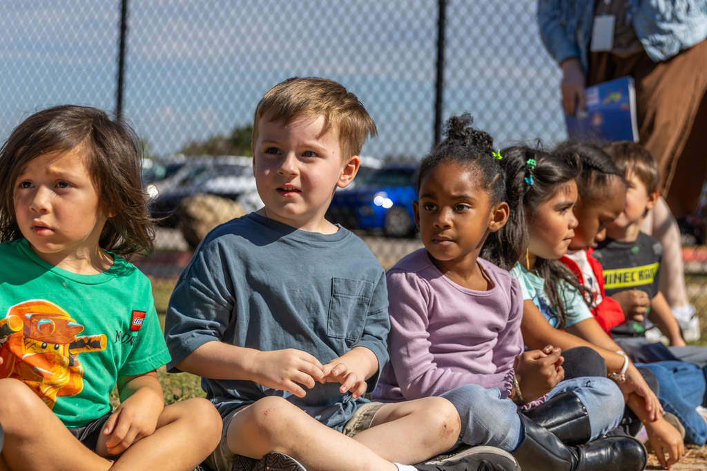A row of Pre-K students sit cross-legged outdoors on the grass, looking attentively toward a storytime presentation.