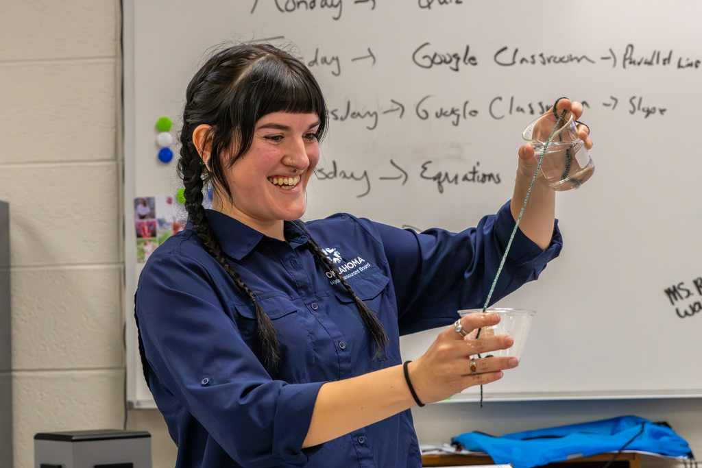 A smiling educator demonstrates a water activity by holding a small glass container above a plastic cup, with a cord extending between them.