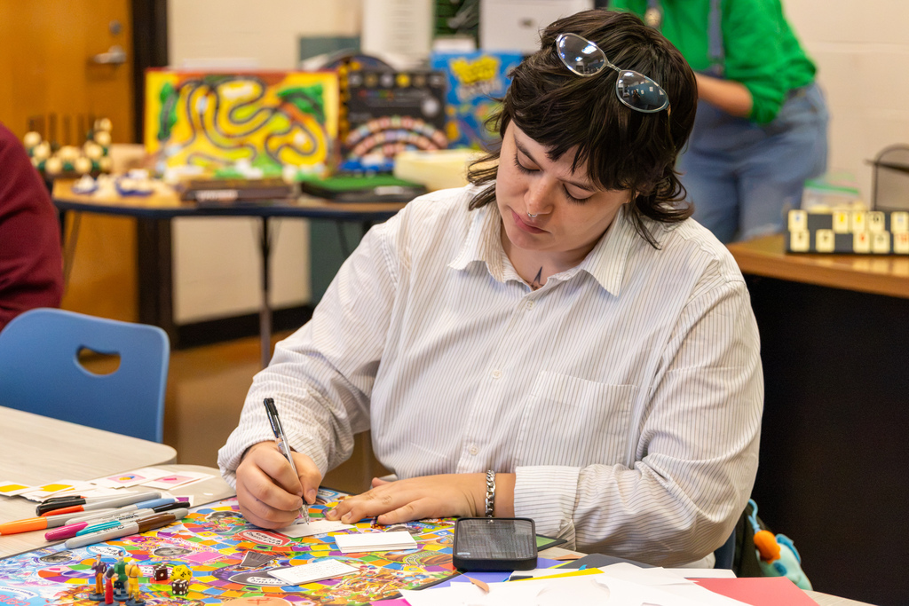 An educator writes on a small card at a table covered with colorful board games, dice, game pieces, markers, and instructional materials.