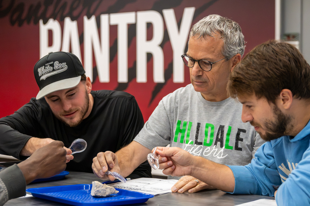 Three educators examine a rock sample on a blue tray, using handheld magnifying glasses during a collaborative geology activity.