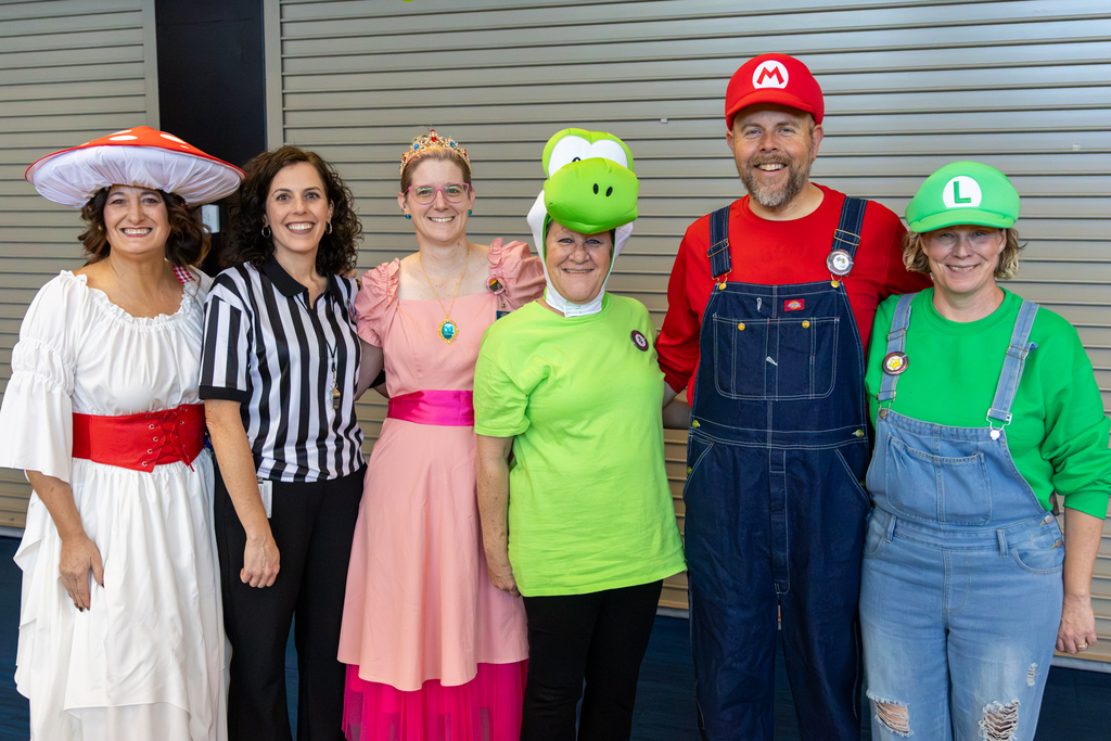 Six Putnam City educators pose together in Mario-themed costumes, including Toad, Princess Peach, Yoshi, Mario, and Luigi, alongside a person dressed as a referee.