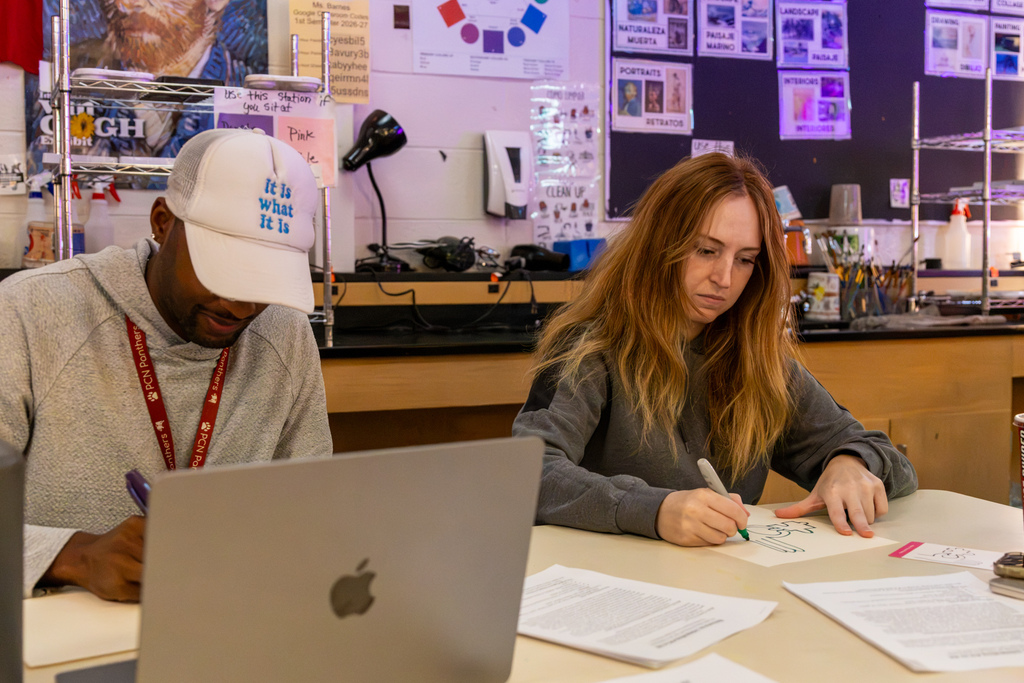 Two educators work independently at a classroom table, one using a laptop while the other draws on paper with a marker.