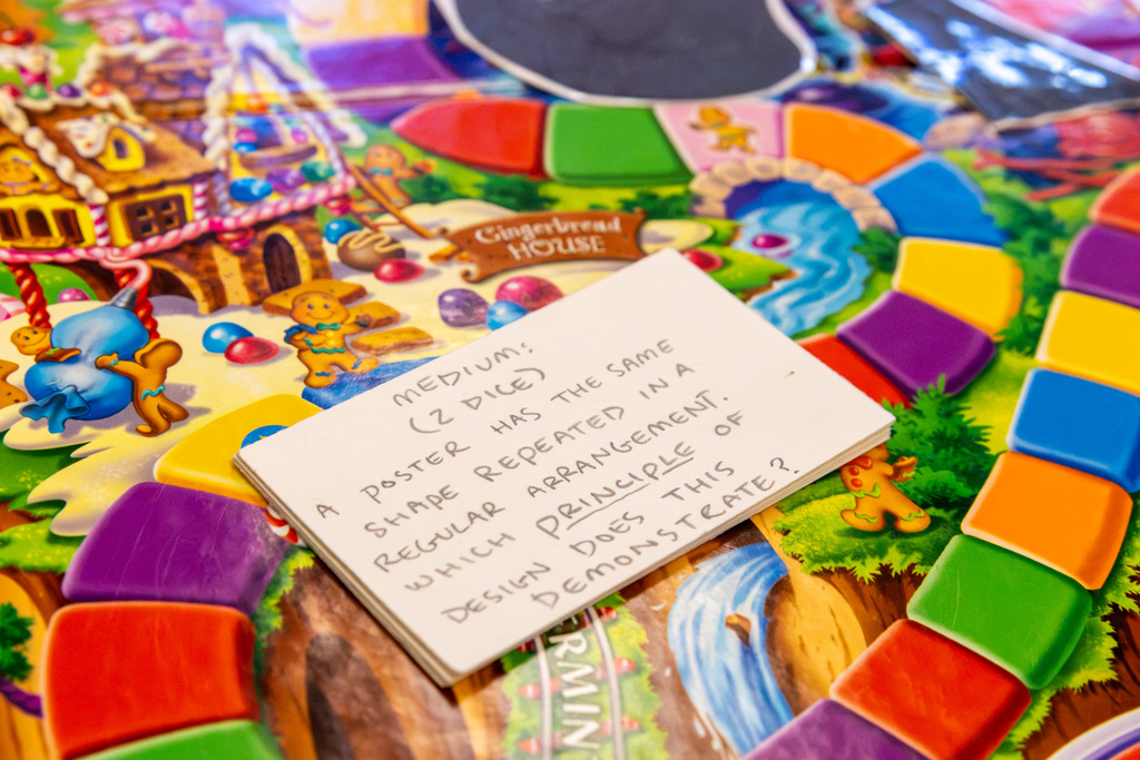 A handwritten question card rests on a colorful Candy Land game board, asking how a poster could be incorporated into a game without changing its design.