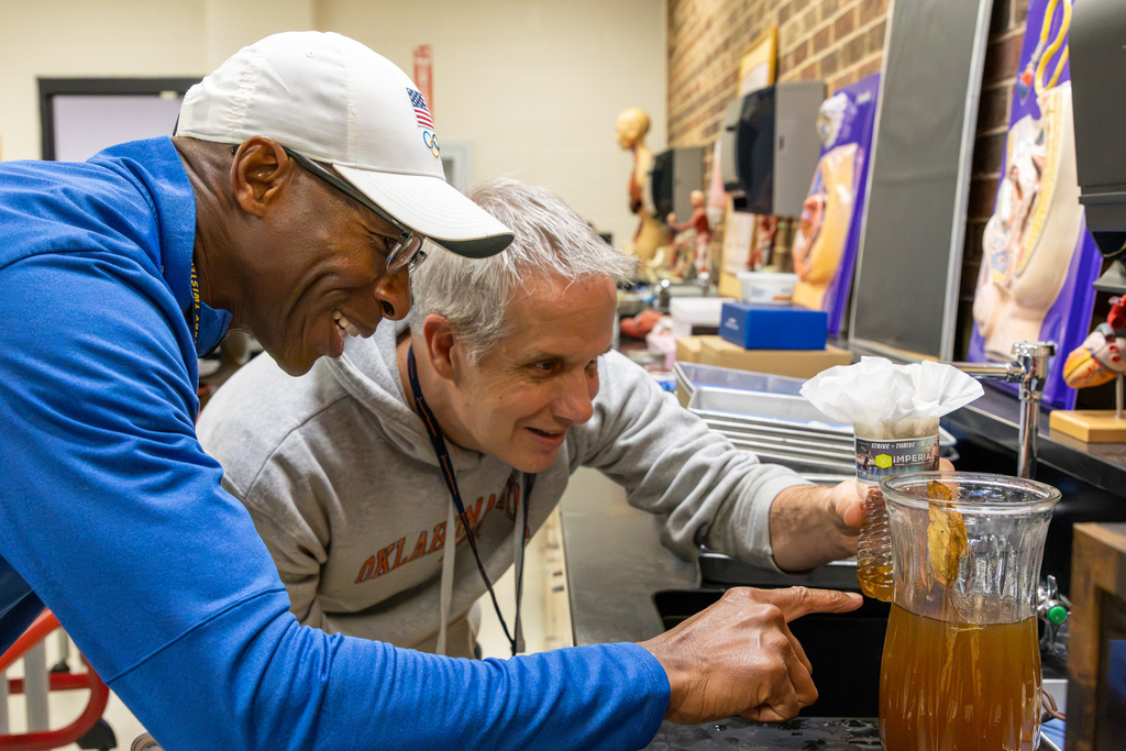 Two educators lean over a classroom counter to closely examine a water-filled pitcher containing materials from a science experiment.