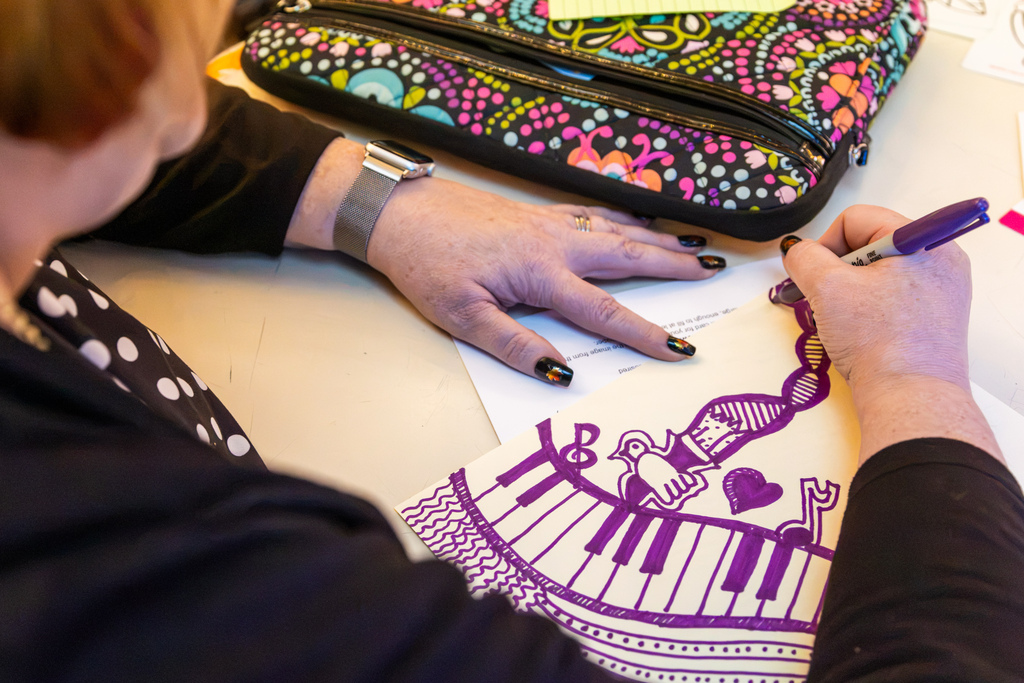 An educator uses a purple marker to draw a colorful illustration featuring a piano keyboard, musical notes, and hearts during a hands-on art workshop.