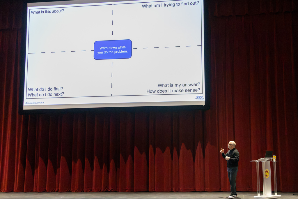 Mathematics educator John SanGiovanni speaks onstage beside a large projected slide showing questions designed to help students explain their mathematical thinking.