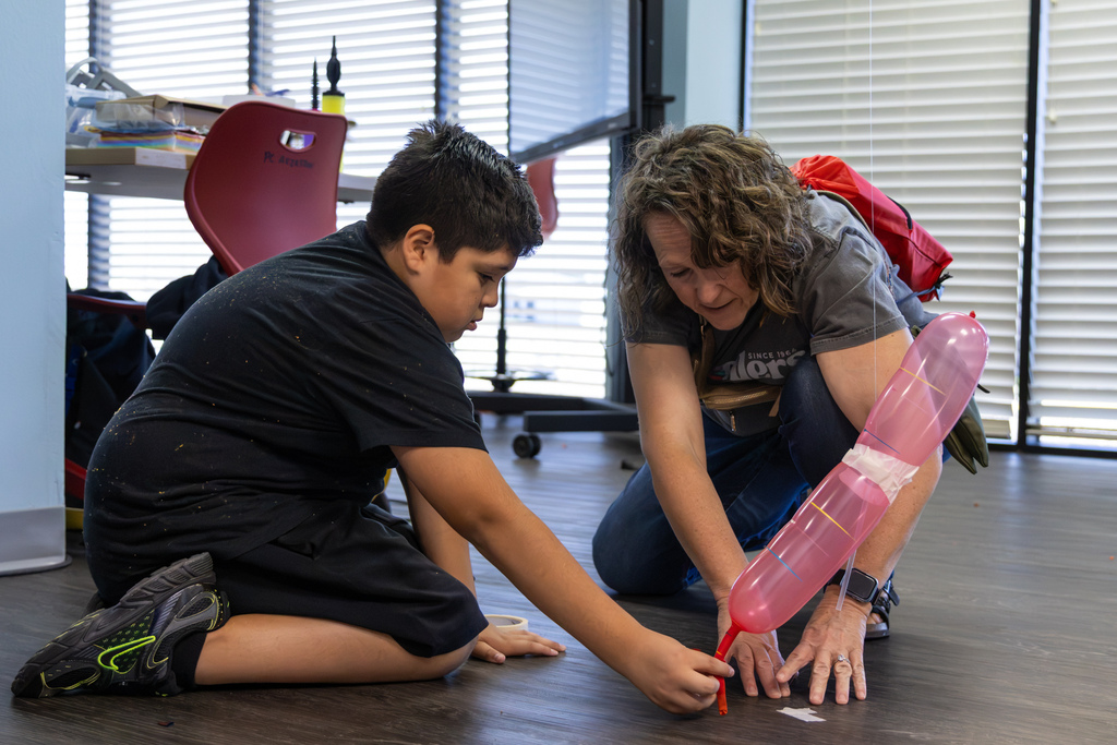 A student and adult work together on the floor, securing a pink balloon rocket setup on a guided line.