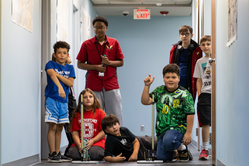 A group of students and high school mentors gather in a hallway set up with vertical launch tubes for a projectile physics experiment.