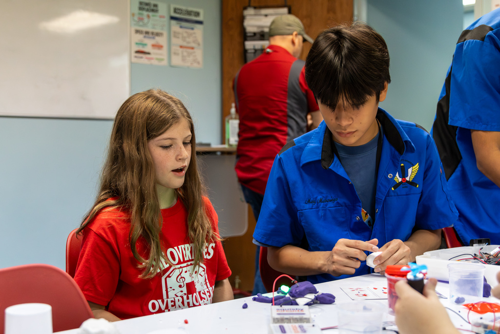 An Overholser Elementary student sits alongside a high school Aviation student at a worktable as they examine a electrical circuit activity with modeling clay and lights.