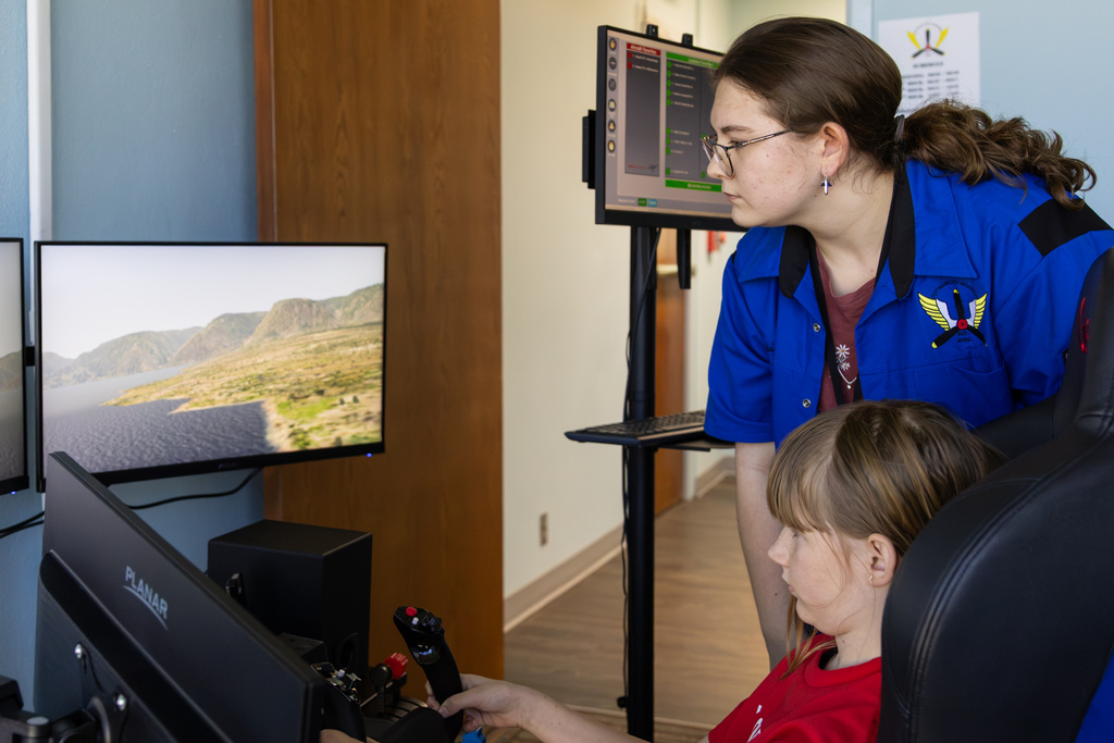 A high school Aviation student leaning over a flight simulator chair to guide a young elementary student through a flight path displayed on a desktop screen.