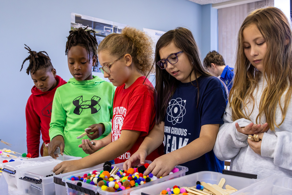 A group of elementary students stand along a table sorting through craft supplies like pom-poms and craft sticks for a STEM activity.