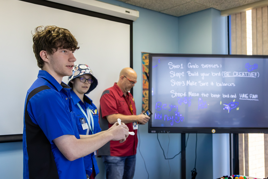 A high school Aviation student stands near an interactive board, holding up a demonstration object while presenting steps for a hands-on STEM project.