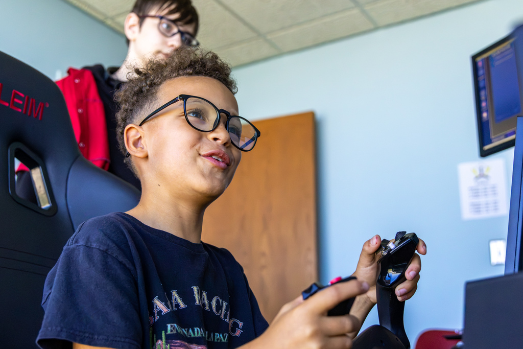A young boy wearing glasses grins enthusiastically as he grips a flight simulator yoke inside an aviation classroom