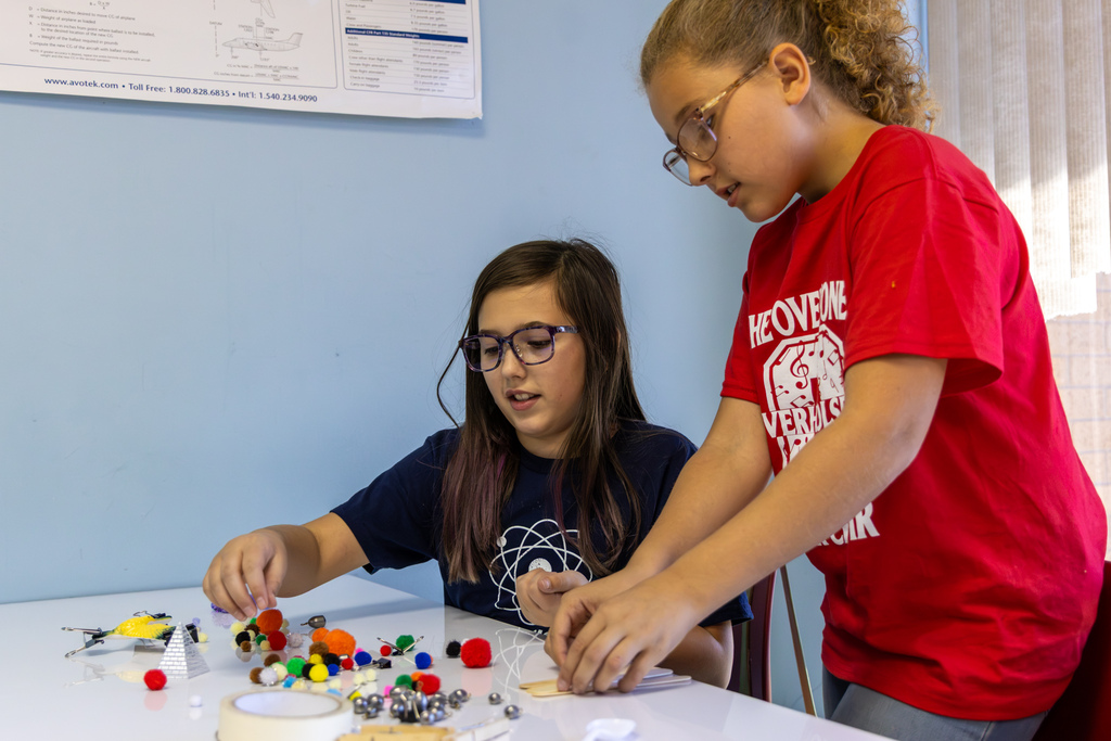 Two students collaborate at a table filled with craft materials to build a balanced bird structure.