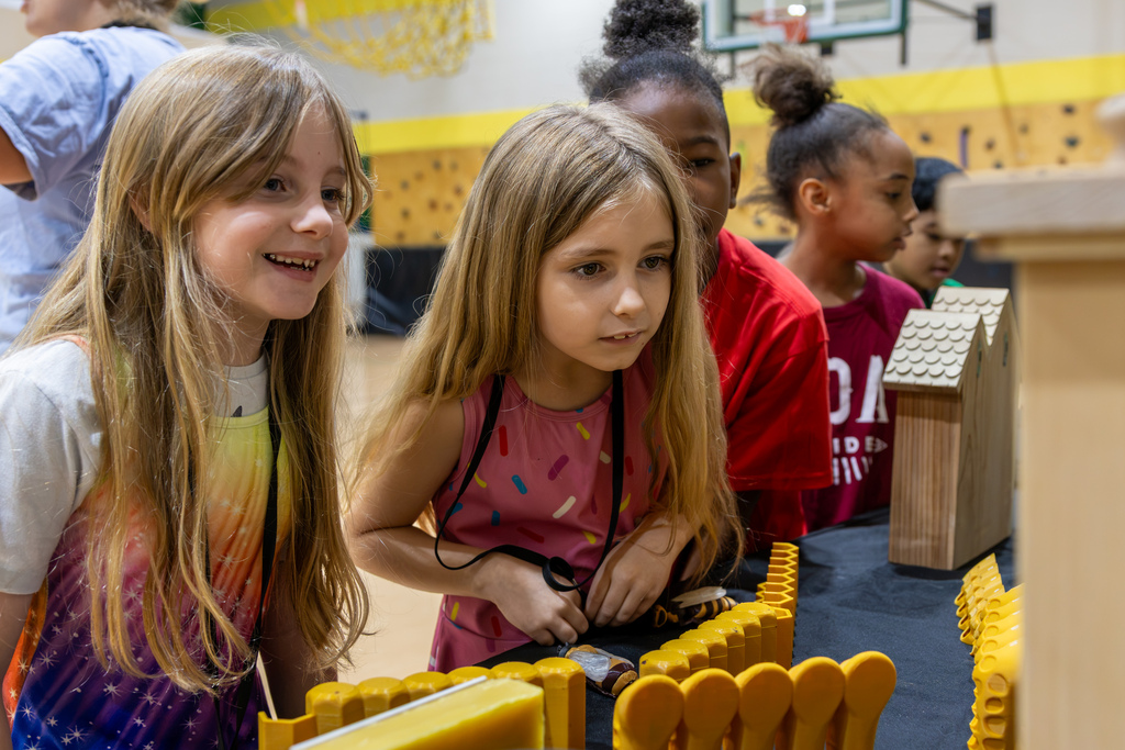 Two elementary girls lean forward with wide smiles to inspect honeycomb models up close.