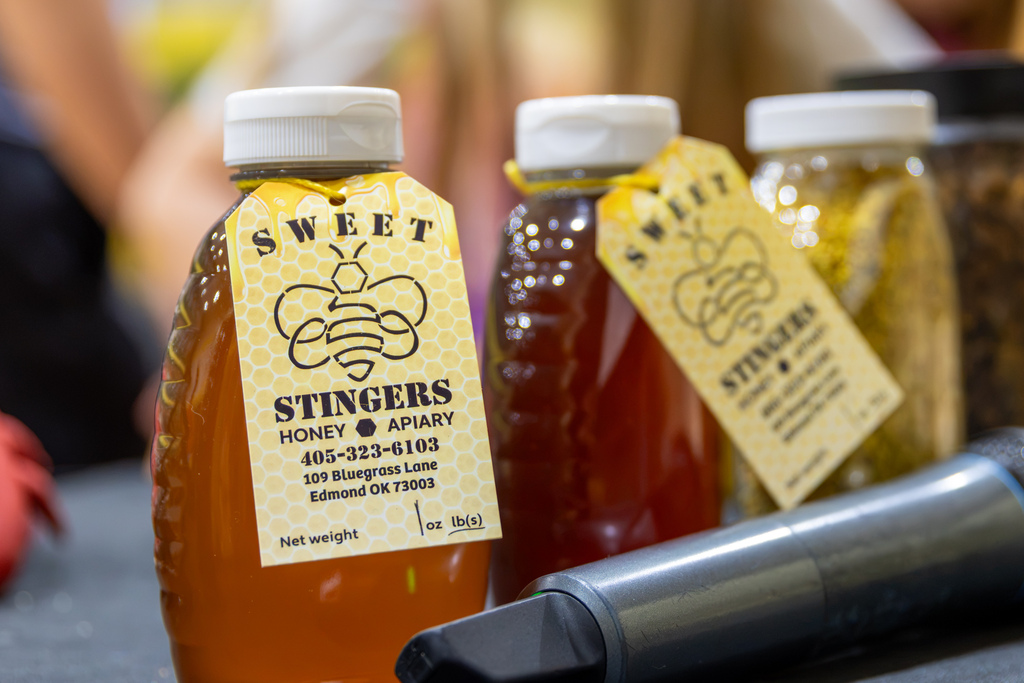 Bottles of Sweet Stingers raw honey with yellow bee-themed labels sitting next to a presenter's microphone on a table.