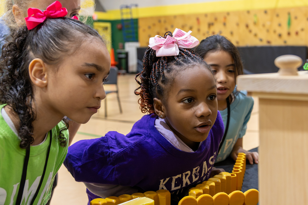Three elementary girls watch intently as a demonstrator explains bee anatomy and honeycomb structures.